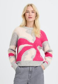 Femme blonde portant un pull abstrait gris é, rose et crème, les mains dans les poches de jeans gris taille haute, sur fond blanc.