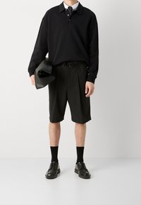 Musta pitkähihaisen poolopaidan, jossa on kolme nappia, yhdistelmä polvimittaisiin mustiin shortseihin ja mustiin kenkiin. Yhdellä kädellä pidetään nahkalaukkua kainalossa.