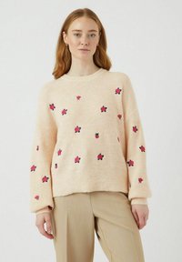 Svijetlo ružičasti pleteni pulover s crvenim i crnim cvjetnim vezom. Ima rebraste manžete i okrugli izrez. Mekana tekstura, opušteni kroj.