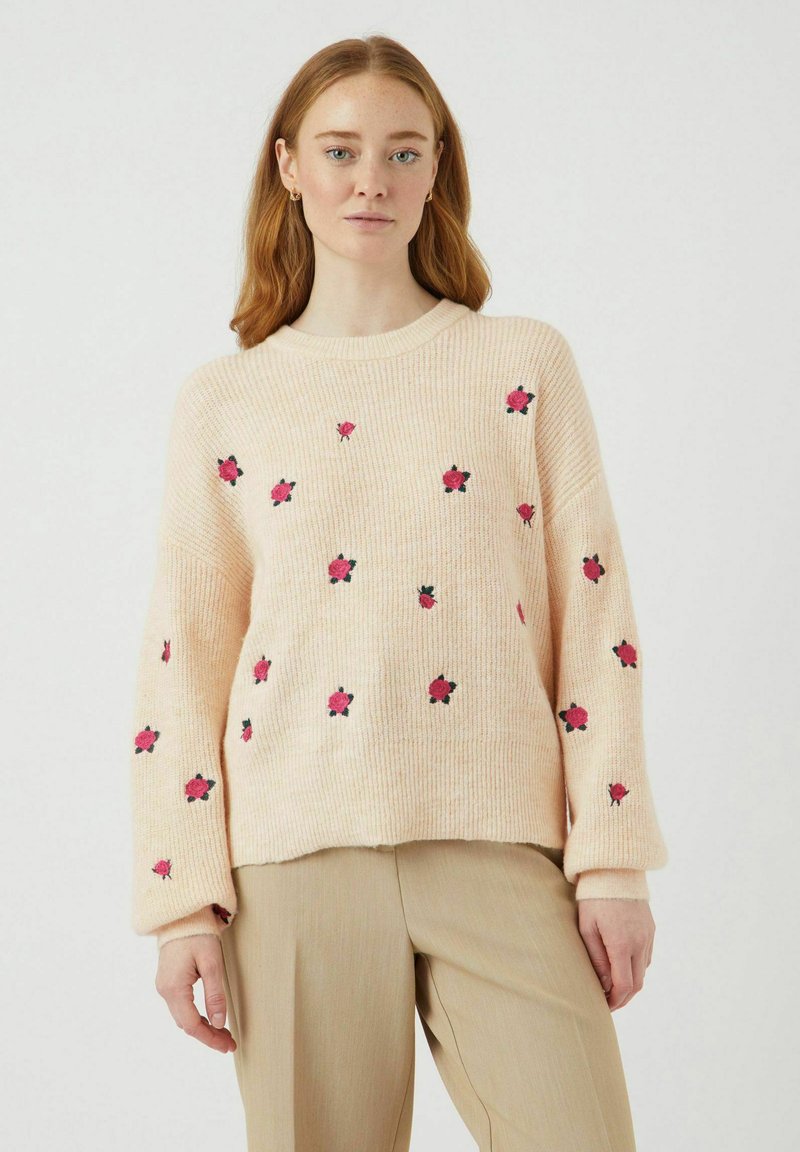 Svijetlo ružičasti pleteni pulover s crvenim i crnim cvjetnim vezom. Ima rebraste manžete i okrugli izrez. Mekana tekstura, opušteni kroj.