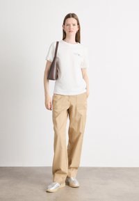 T-shirt bianco in cotone con logo stampato, pantaloni cargo color khaki con tasche e sneakers argento. Borsa marrone dal design elegante e minimalista.