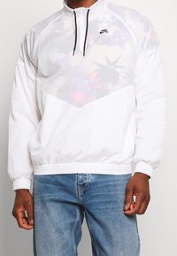 Giacca Nike SB bianca con mezza cerniera, tessuto leggero, presenta un pannello traslucido sul petto con motivo floreale colorato, polsini e orlo elasticizzati.
