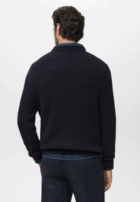Suéter de punto de canalé en color azul marino, con una textura tejida, que presenta un cuello alto y mangas largas, combinado con una camisa de mezclilla.