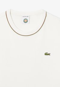 T-shirt bianco in cotone con collo rotondo, caratterizzato da un colletto rifinito in verde e rosso e un piccolo logo di coccodrillo ricamato in verde.