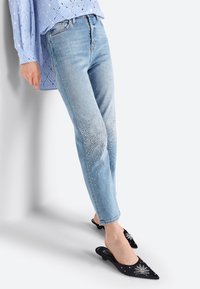 Jasnoniebieskie spodnie dżinsowe o slim fit, zdobione rozproszonymi kryształowymi detalami od kolana do kostki. W zestawieniu z czarnymi ostrymi mule z dekoracyjnymi akcentami.