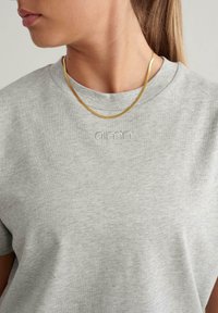 Graues Baumwoll-T-Shirt mit Rundhalsausschnitt und kurzärmeligen. Mit dezentem "aimn"-Stick auf der Brust. Getragen mit einer dünnen goldenen Kette.
