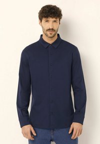 Camicia a maniche lunghe color navy con chiusura a bottoni, colletto a punta e orlo dritto. Il tessuto sembra lucido e liscio.