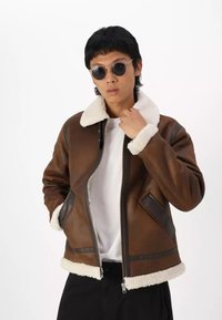 Giacca in pelle marrone con colletto in pelliccia, chiusura a zip frontale e tasche anteriori; presenta un tessuto testurizzato e accenti scuri a contrasto.