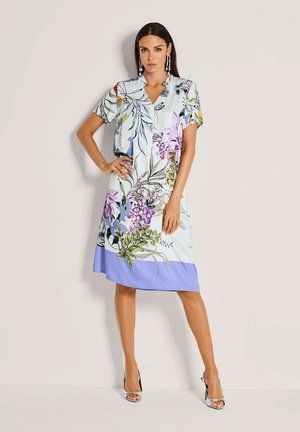 Bloemenjurk met korte mouwen, een V-hals en een pastelblauwe onderkant. De stof toont diverse plantenpatronen in paars en groen.