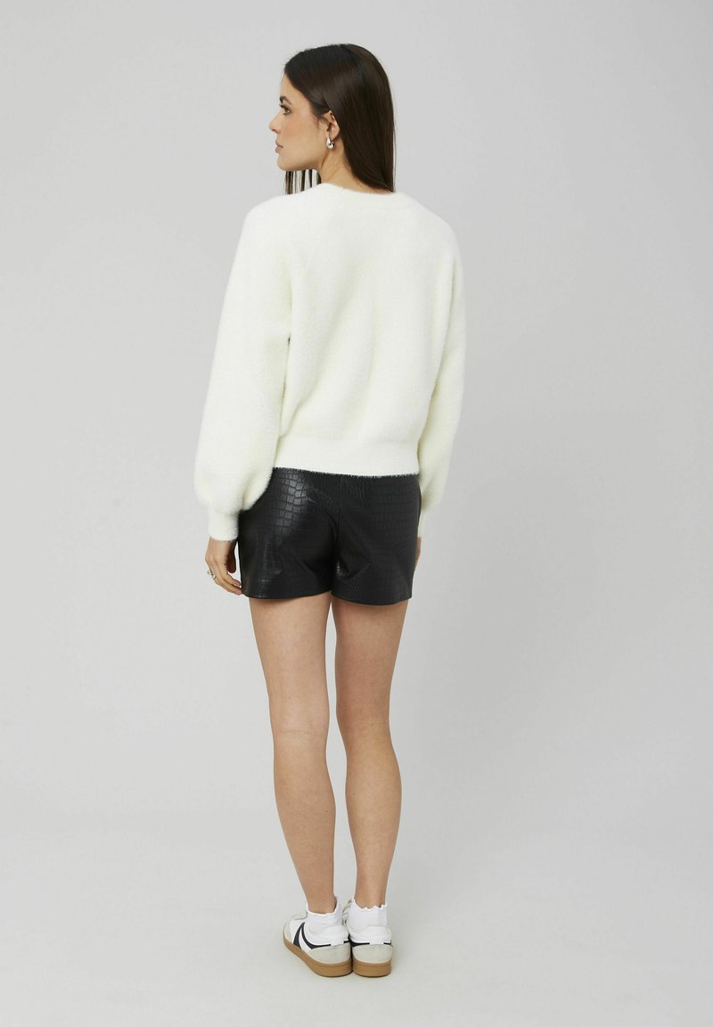 Pull en laine blanc et duveteux avec un col rond associé à un short noir texturé. La modèle montre le dos, portant des baskets blanches.