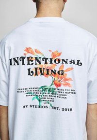 T-shirt alb din bumbac, cu un design floral în nuanțe de portocaliu și verde, având text negru îngroșat care spune "VIAȚĂ INTENȚIONATĂ" și un text suplimentar dedesubt.