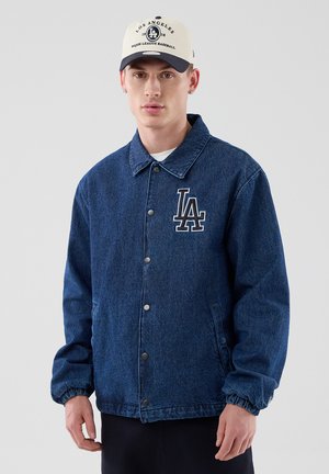 Młody mężczyzna w niebieskiej dżinsowej kurtce z logo LA oraz białą i granatową czapką baseballową Los Angeles, stojący na jednolitym tle.