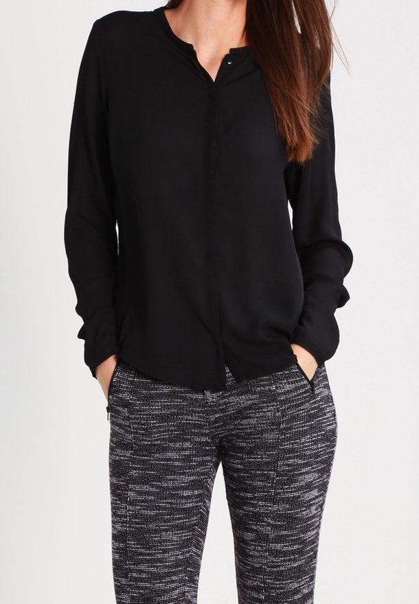 Femme portant un chemisier noir à manches longues, rentré dans un pantalon à motif noir et blanc, les mains dans les poches.
