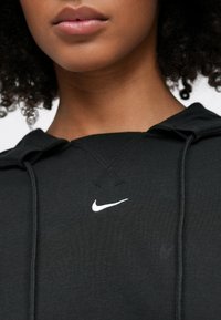Közeli kép egy személy alsó arcáról és nyakáról, aki fekete kapucnis pulóvert visel, a mellkas közepén fehér Nike logóval.