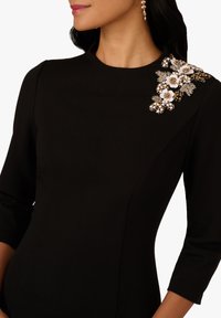 Robe noire à manches trois-quarts, ornée de détails floraux embellis sur l'épaule, fabriquée en tissu structuré et lisse.