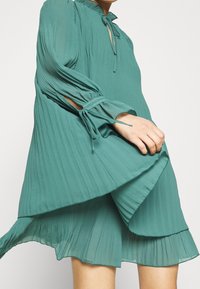 Miętowa plisowana sukienka z lekkiego materiału, z wysokim dekoltem, dzwonowymi rękawami z wiązaniami oraz rozszerzonym dołem.