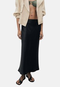 Jupe maxi en satin noir avec une texture lisse et un ourlet légèrement évasé. Associée à une veste courte beige et des sandales décontractées.