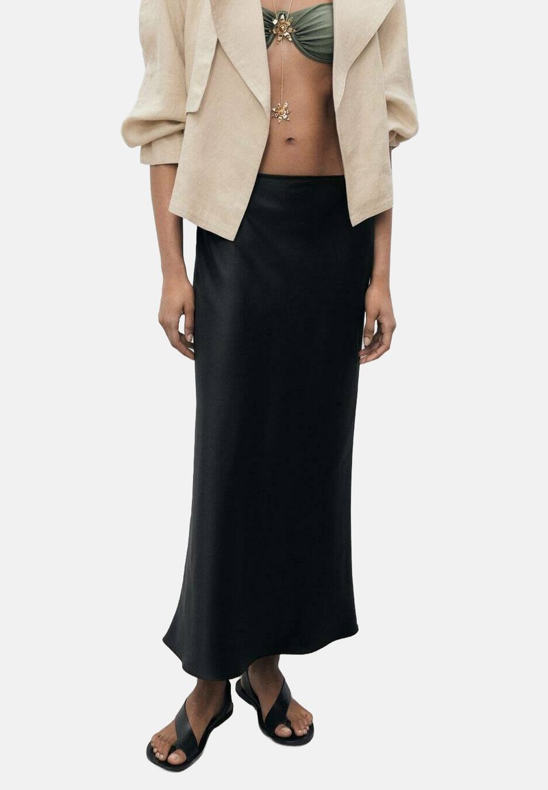 Jupe maxi en satin noir avec une texture lisse et un ourlet légèrement évasé. Associée à une veste courte beige et des sandales décontractées.