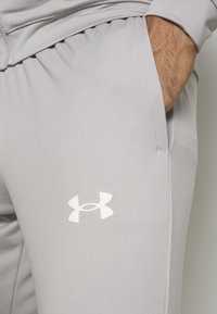 Pantalon de sport gris clair avec une main dans la poche droite et un logo blanc Under Armour sur la cuisse.