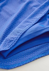 Piezas de tela atlética azul superpuestas: una con una superficie texturizada y similar a una malla, y otra lisa, ambas mostrando un color vibrante y detalles de costura.