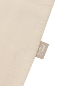 Tissu de muslin beige avec une surface douce et texturée. Comprend une petite étiquette grise avec un logo, positionnée sur le bord.