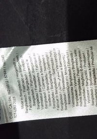 Étiquette de vêtement indiquant « Fabriqué au Bangladesh » imprimé en plusieurs langues sur un tissu blanc sur un fond sombre.