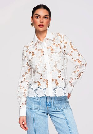 Femme portant une chemise blanche à manches longues en dentelle florale avec un col et un jean taille haute bleu clair, posant devant un fond uni.