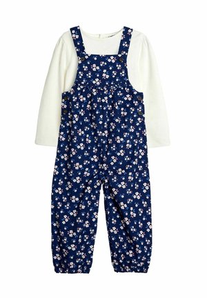 Peto floral azul marino con flores rosas y blancas, con cierre de botón y puños elásticos, superpuesto sobre una camiseta de manga larga blanca.