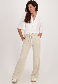 Vrouw glimlachend, gekleed in een witte blouse met een strikhals en brede beige broek met een strik, met handen in de zakken, staand tegen een effen achtergrond.