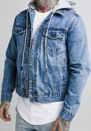 Hombre que lleva chaqueta vaquera azul con capucha gris sobre camisa blanca, con tatuajes visibles en el cuello y las manos, de pie frente a un fondo claro.