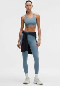 Sujetador deportivo azul claro y leggings a juego, con un diseño ajustado, textura suave y una camiseta negra anudada en la cintura. Zapatillas blancas con detalles.