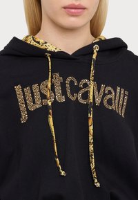 Primo piano di una persona che indossa un hoodie nero con lacci gialli a fantasia e il logo dorato con borchie "Just Cavalli" sul petto.