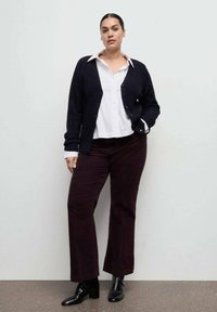 Femeie stând sprijinită de un perete simplu, purtând o cămașă albă cu guler, un cardigan negru, pantaloni întunecați și bocanci negri de gleznă cu toc.