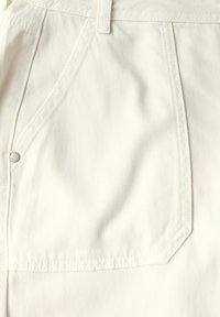 Detail bílé denimové látky s blízkým záběrem na kapsu s prošíváním a malým kovovým knoflíkem. Hladká textura a strukturovaný tvar.