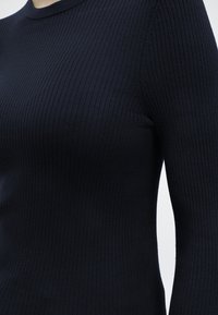 Κοντινό πλάνο ενός ατόμου που φορά στενή μαύρη ριπ κοντομάνικη μπλούζα με στρογγυλή λαιμόκοψη, πάνω σε απλό φόντο.