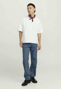 Camisa branca de manga curta com decote redondo, combinada com jeans de ganga azul. Uma bandana vermelha está atada ao pescoço. Sapatos pretos completam o visual.