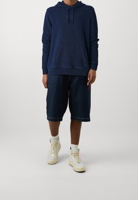 Felpa navy con tasca frontale e logo, abbinata a pantaloni corti in denim a ginocchio con un accento rosso. Sneakers bianche alte completano il look.