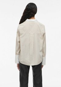 Gestreepte button-up shirt in licht crèmekleur met subtiele roze lijnen. Heeft lange mouwen, witte manchetten en een schouderdetail op de achterkant.