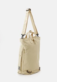 Béžový nylonový batoh s nastavitelným ramenním popruhem, horními rukojeťmi, boční kapsou na zip a plochým dnem pro stabilitu. Minimalistický design bez vzorů.