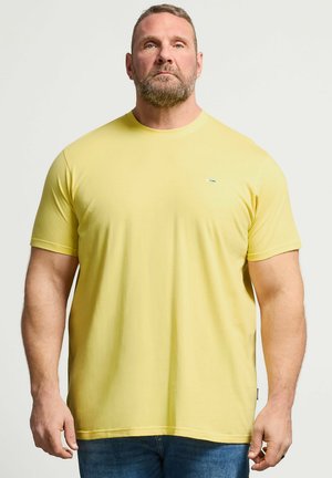 Brodaty mężczyzna w gładkiej żółtej koszulce z krótkim rękawem i niebieskich dżinsach, stojący na neutralnym tle, patrzący prosto.