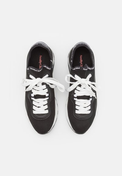Zwarte sneakers met witte veters, textuur bovenkant, logo-accenten op de tong en een gewatteerde witte zool, gezien van recht boven.