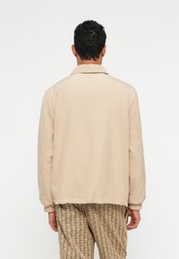 Beige jaka ar apkakli, elastīgām piedurknēm un apakšmali. Gluda tekstūra, redzama aizmugurējā skata. Savienota ar rakstainām beigu biksēm.