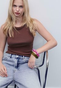 Canottiera a costine marrone, jeans di denim in denim chiaro a vita alta e due bracciali spessi - uno verde e uno rosa - sul polso destro.