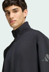 Čierna zipsová bunda s vysokým golierom, vyrobená z jemnej tkaniny. Obsahuje matne čierne trojuholníkové logo na ľavom rukáve a minimalistický dizajn.