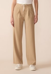 Beige wijde broek met een trekkoord in de taille en geplooide voorkant, gemaakt van lichte stof, gecombineerd met witte sneakers.