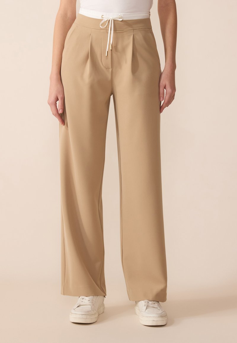Beige wijde broek met een trekkoord in de taille en geplooide voorkant, gemaakt van lichte stof, gecombineerd met witte sneakers.