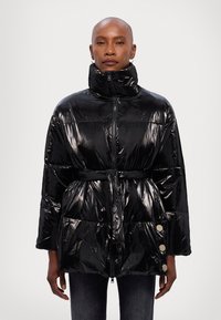 Chaqueta de plumas negra con un acabado brillante, cuello alto, cintura ceñida con cinturón y detalles de botones en los bolsillos. Presenta un cierre delantero de cremallera y mangas anchas.