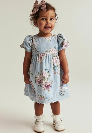 Himmelblaues Blumenkleid mit Puffärmeln, Spitzenrand und rosa, lila sowie blauen Blumen. Kombiniert mit weißen Schuhen und Spitzenstrümpfen.