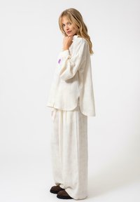Completo in lino bianco con una maglietta a maniche lunghe e svolazzante, dotata di uno spacco laterale, e pantaloni a gamba larga con un motivo sottile. Calzature marroni completano il look.