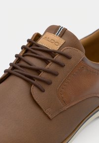 Chaussures en cuir synthétique marron avec une finition texturée, dotées de lacets marron, d'une patch en cuir tan étiqueté "ALDO" et d'un accent rayé contrastant.